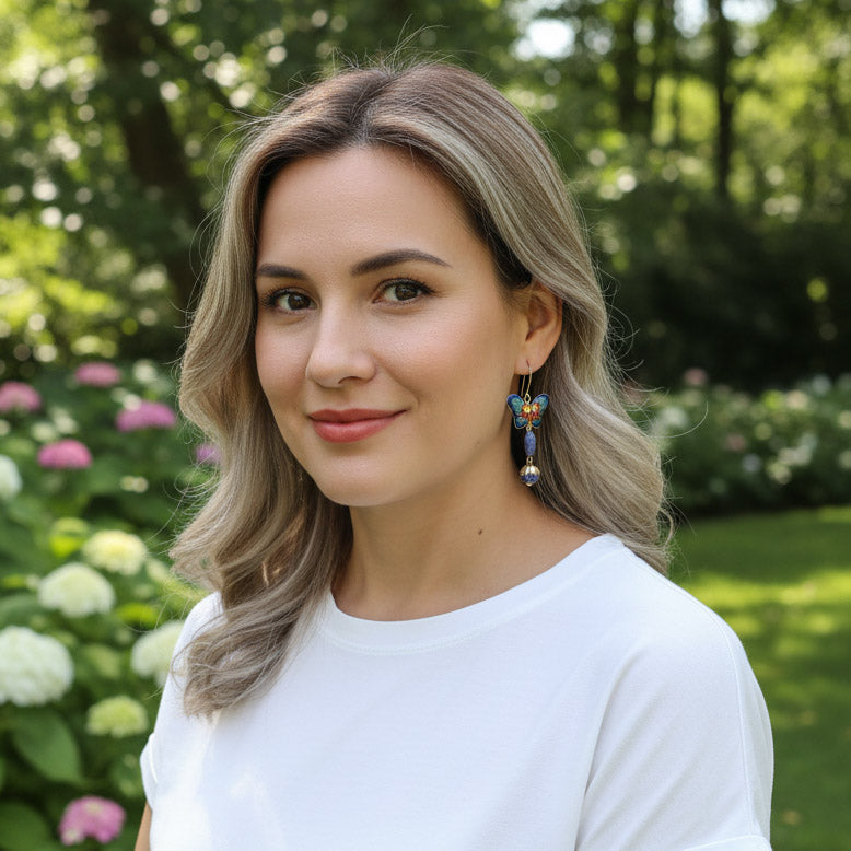 Model wearing gold-plated cloisonné butterfly drop earrings with blue lapis beads in garden sunlight.