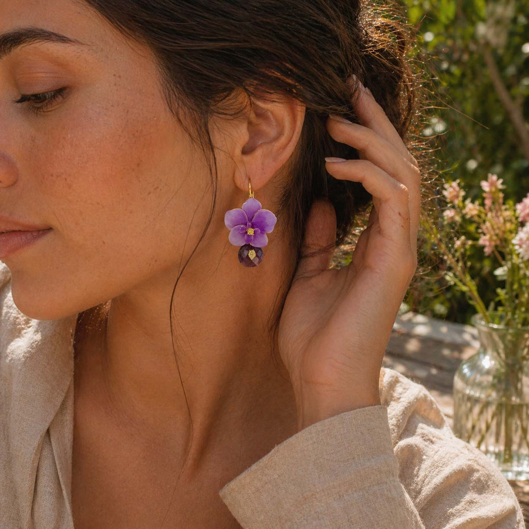 Woman wearing a purple orchid flower earring outdoors with greenery in the background