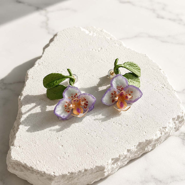 Purple orchid enamel stud earrings displayed on a textured white stone surface, featuring hand-painted petals, green resin leaves and natural stone chip accents.