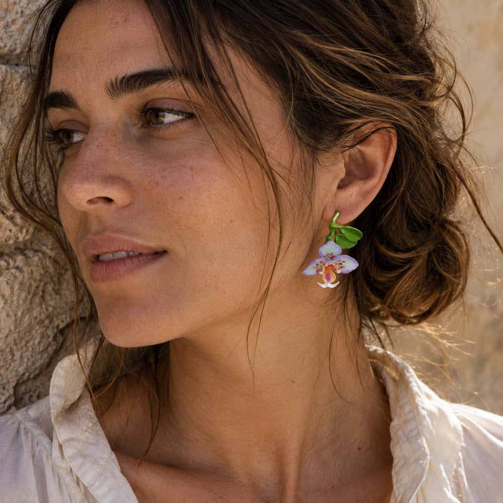 Woman wearing handcrafted purple orchid enamel stud earrings with sculpted green leaves and botanical floral design.