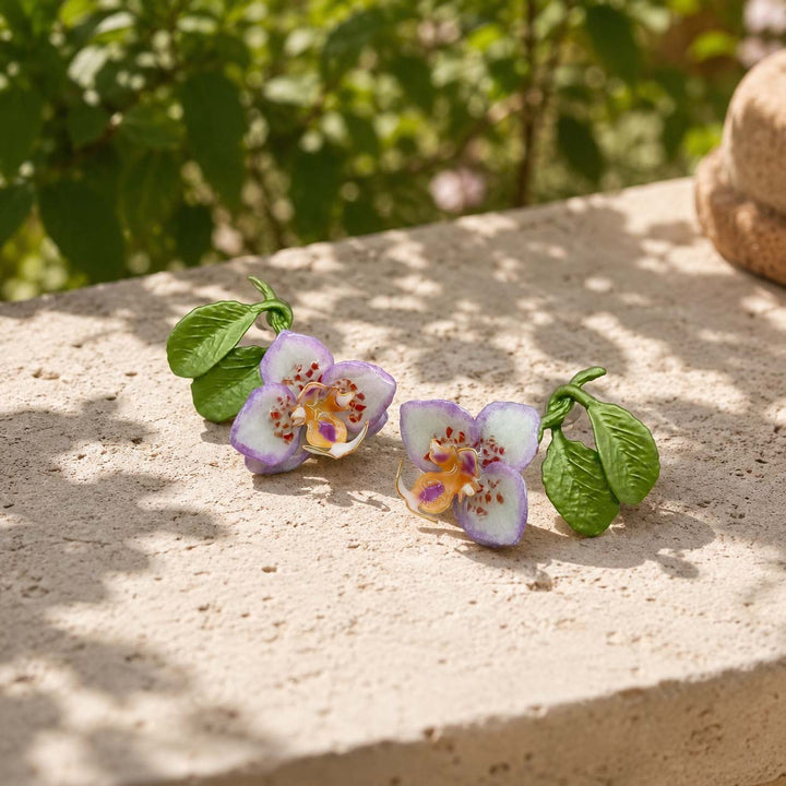 Hand-painted purple orchid enamel stud earrings styled on a natural stone surface in a sunlit garden, featuring green resin leaves and semi-precious stone chip accents.