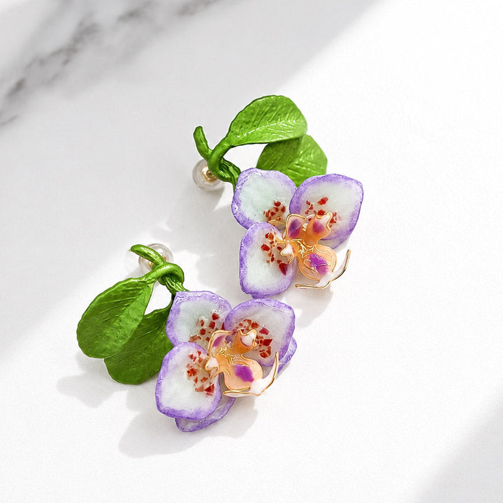 Hand-painted purple orchid enamel stud earrings with sculpted green resin leaves and semi-precious stone chip accents on a white marble background.
