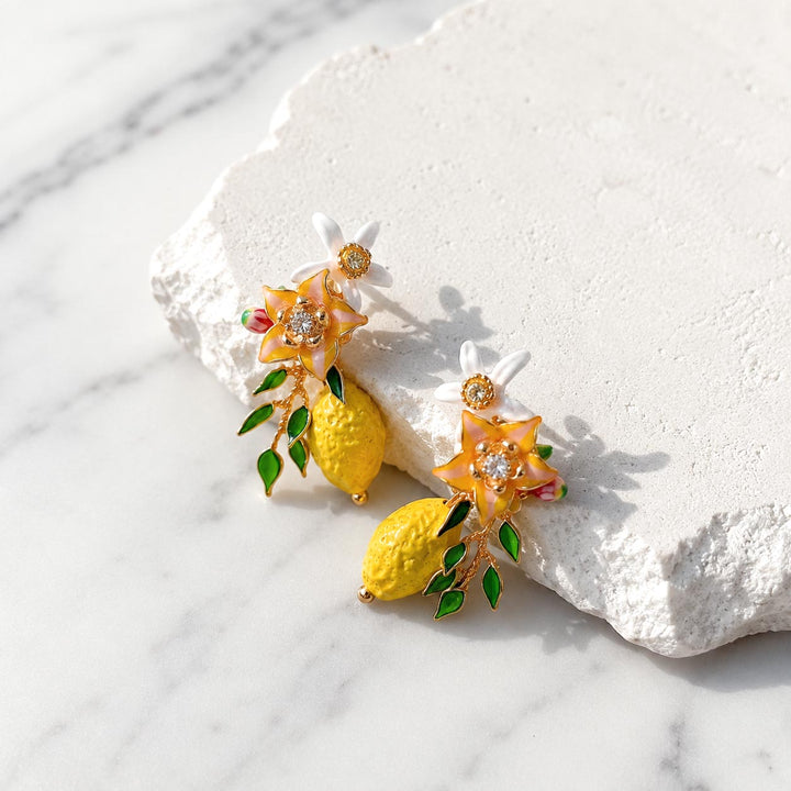Hand-painted lemon blossom stud earrings displayed on a textured white stone surface with enamel flowers, resin blossoms and lemon charms.