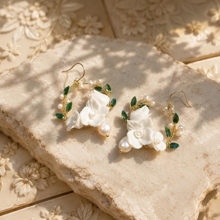 White porcelain flower hoop earrings with hand-painted green enamel leaves on a textured stone surface.