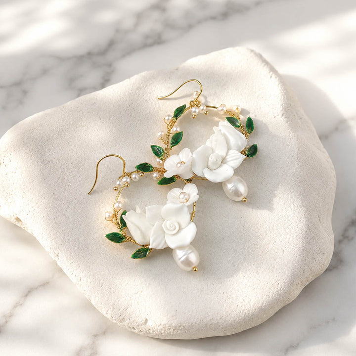 White porcelain flower hoop earrings with hand-painted green enamel leaves on a marble surface
