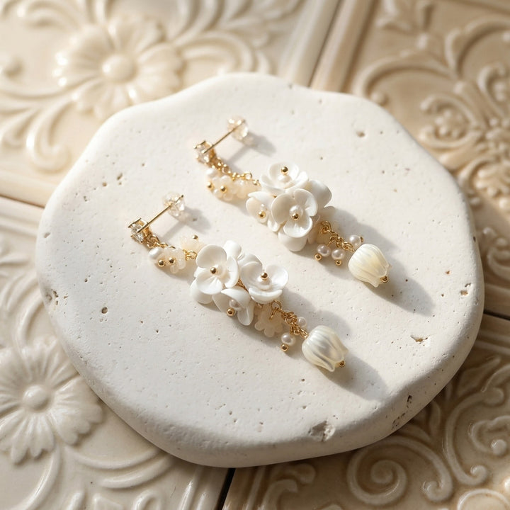 Porcelain flower and mother-of-pearl tulip drop earrings with CZ studs and freshwater pearls on pale textured stone.