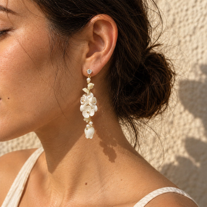 Model wearing white porcelain floral drop earrings with mother-of-pearl tulips and pearl accents outdoors against a pale stone wall.