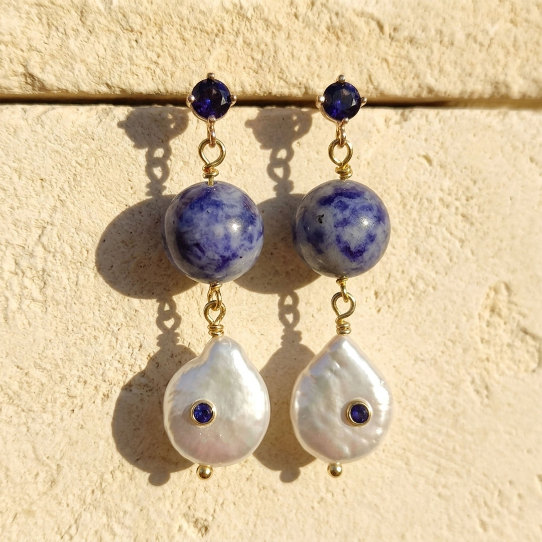 A pair of gold-tone dangle earrings featuring sodalite beads, a sapphire color CZ stone at the top, and a larger white baroque pearl with a tiny dark blue stone at the bottom, displayed against a textured beige wall.