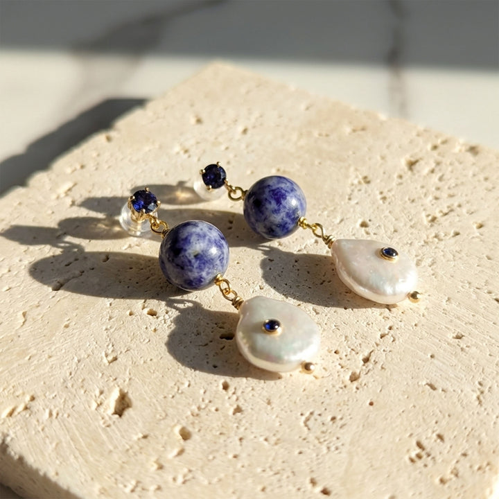 A pair of bohemian-style dangle earrings resting on a textured beige stone slab. The earrings feature round natural sodalite bead, irregular white baroque pearls, and small dark blue gemstone set in gold-tone metal.