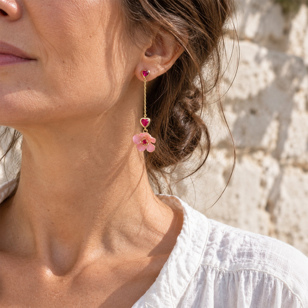 A close-up lifestyle shot of a woman wearing asymmetric Sakura heart drop earrings with pink CZ stones, set against a blurred stone wall background.