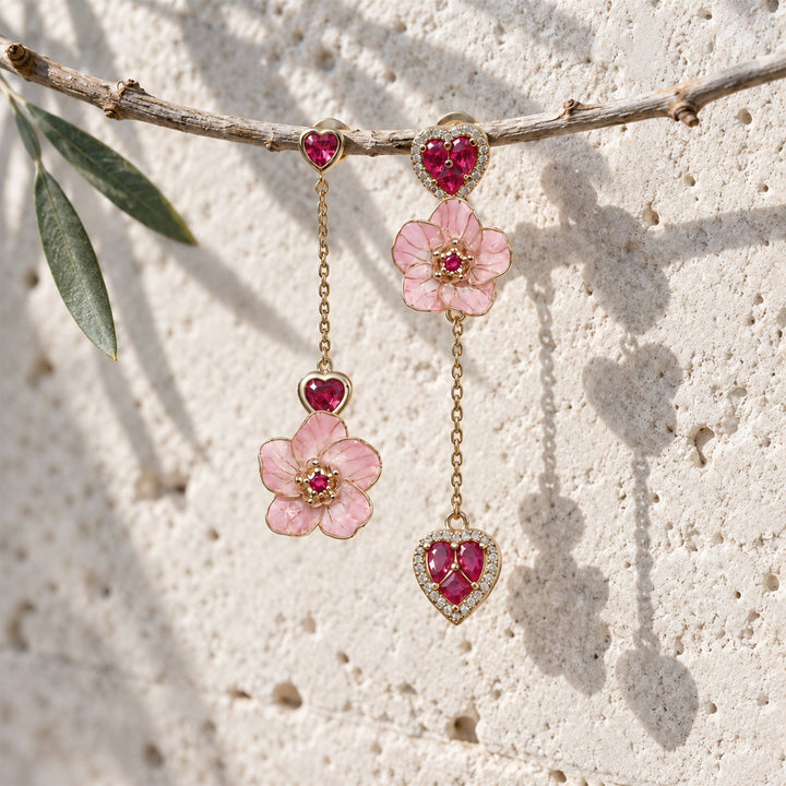A pair of gold dangle earrings with pink cherry blossom flowers and red heart-shaped stones, hanging from a rustic branch against a textured light stone wall.