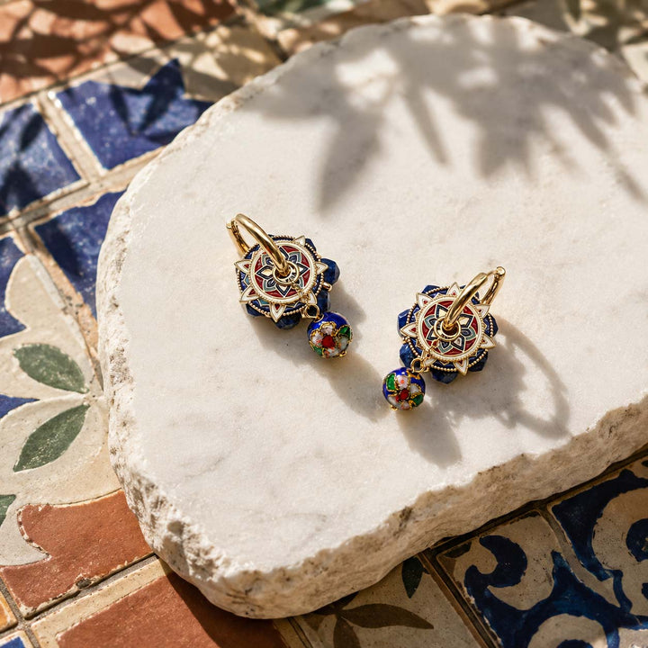 A pair of gold hoop earrings with intricate multicolor enamel floral clusters and dangling cloisonne charms, displayed on a white stone slab over patterned blue and terracotta tiles.
