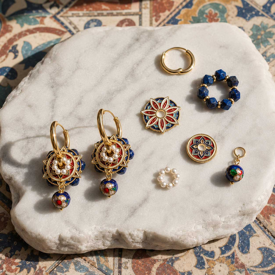 A styled flat-lay of lapis and cloisonné convertible hoop earrings, arranged on a white marble tray over a patterned textile background. The set includes hoop earrings, drop earrings, freshwater pearls rings, and round enamel charms.