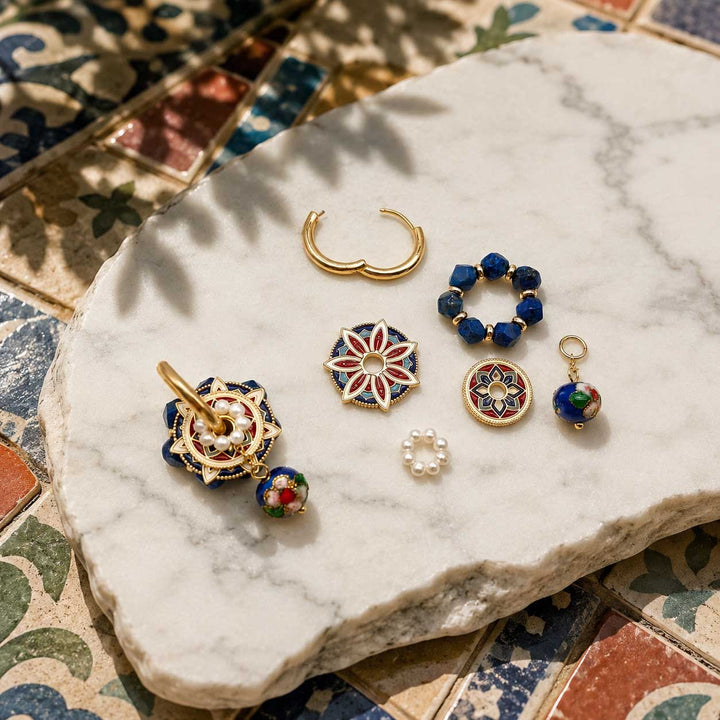 A styled flat-lay of several handmade gold and enamel jewelry pieces arranged on a white marble slab over patterned ceramic tiles. The jewelry includes a crescent hoop, beaded rings, a floral brooch, a small pearl bead, and a decorative earring with multicolor enamel.