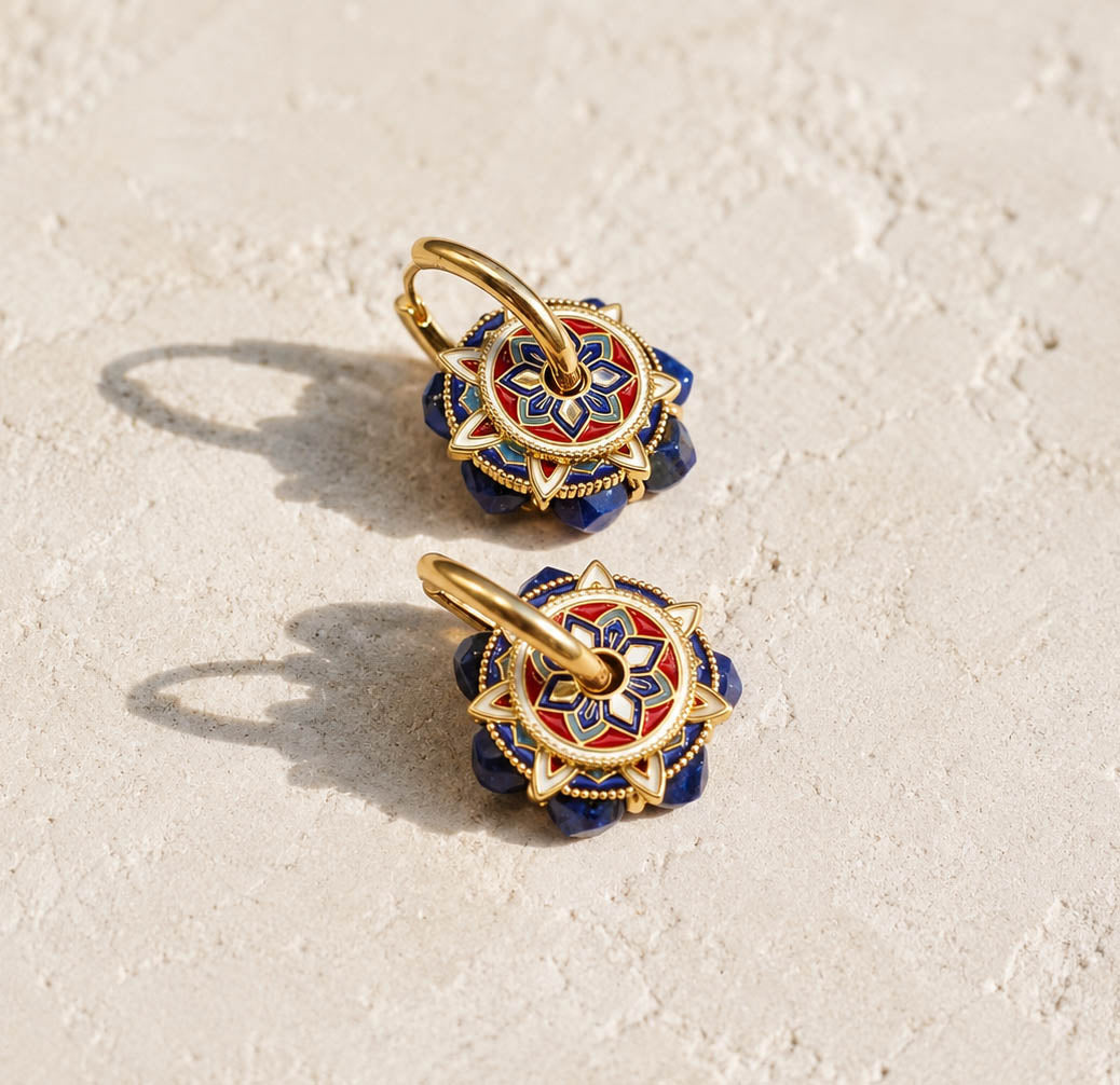 A pair of gold-toned hoop earrings with circular floral charms featuring blue, red, and white enamel detailing, displayed on a textured beige surface.