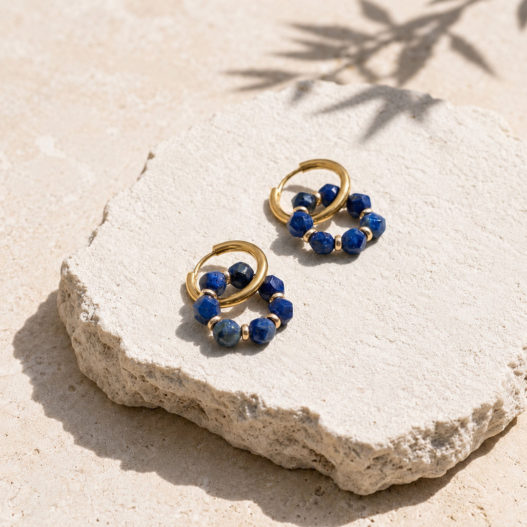 A pair of gold hoop earrings with lapis stone bead clusters, displayed on a textured light beige stone slab outdoors with natural shadows.