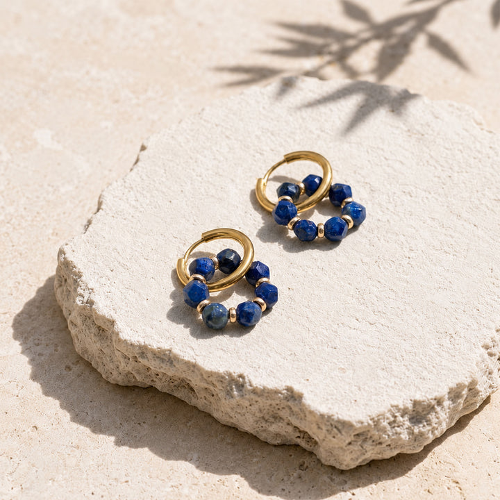 A pair of gold hoop earrings with lapis stone bead clusters, displayed on a textured light beige stone slab outdoors with natural shadows.