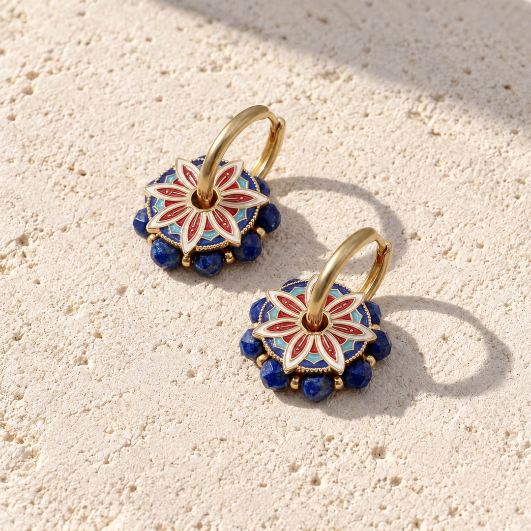 A pair of gold hoop earrings with circular floral pendants featuring red, white, and blue enamel detailing and lapis beads, displayed on a textured beige stone surface.