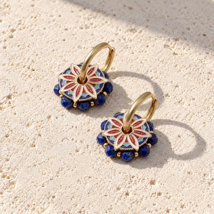 A pair of gold hoop earrings with circular floral pendants featuring red, white, and blue enamel detailing and lapis beads, displayed on a textured beige stone surface.