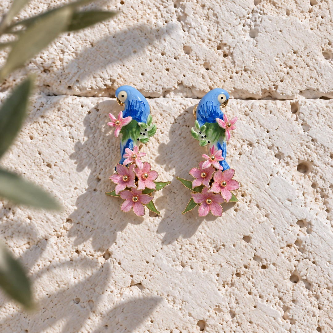 Pair of blue parrot floral drop earrings with pink flowers and green leaves, displayed on a textured light stone surface with soft leaf shadows.