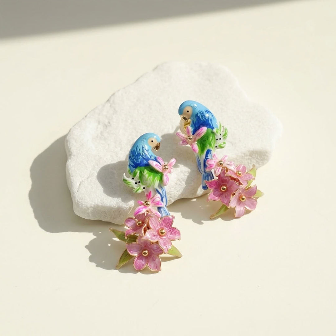 A pair of colorful enamel bird stud earrings featuring blue and green parrots holding pink handcrafted flowers, displayed on a textured white stone against a warm cream background.