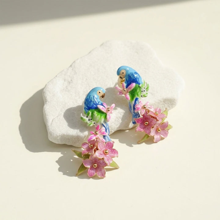 A pair of colorful enamel bird stud earrings featuring blue and green parrots holding pink handcrafted flowers, displayed on a textured white stone against a warm cream background.