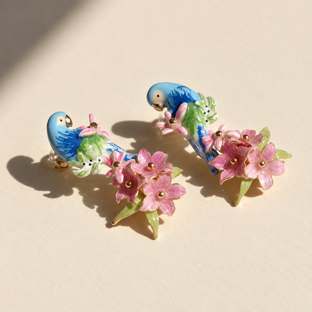 A pair of enamel pin earrings featuring blue and green parrots perched on pink flowers with gold-tone metal details, displayed on a warm beige surface with soft shadows.