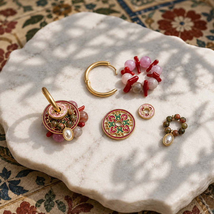 Convertible hoop earring set with pink quartz, unakite jasper, coral accents and hand-painted enamel components arranged on a white marble slab with dappled sunlight. 