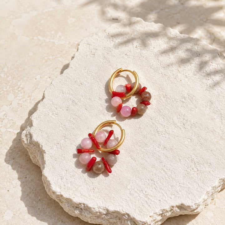A pair of gold hoop earrings with dangling coral and rose quartz beads, displayed on a textured white stone slab.