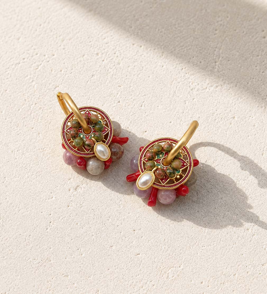 A pair of gold hoop earrings with circular mandala-style beadwork in red, green, and gold, accented by rose quartz, coral, resting on a textured beige surface with soft shadows.