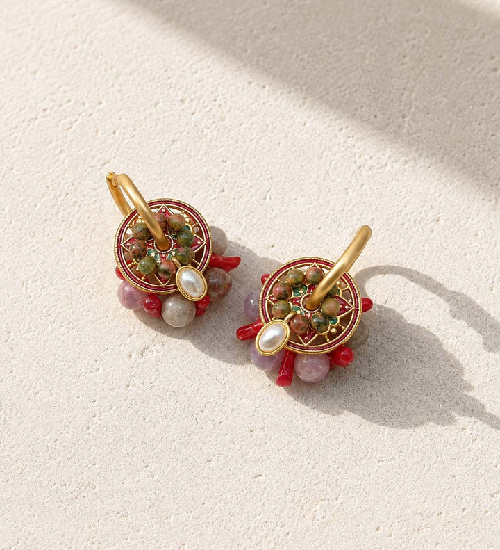 A pair of gold hoop earrings with circular mandala-style beadwork in red, green, and gold, accented by rose quartz, coral, resting on a textured beige surface with soft shadows.
