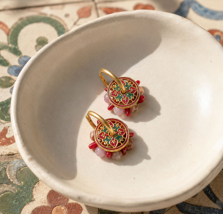 A pair of ornate gold-toned earrings with colorful floral enamel detailing and dangling pink beads, displayed inside a white ceramic bowl on a patterned surface.