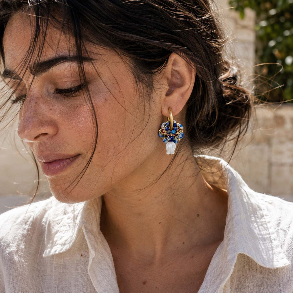 A close-up lifestyle shot of a woman wearing a wearing handcrafted blue porcelain convertible hoop earrings with lapis beads, cloisonné details and carved mother of pearl tulip drops., set against a soft-focus outdoor background.