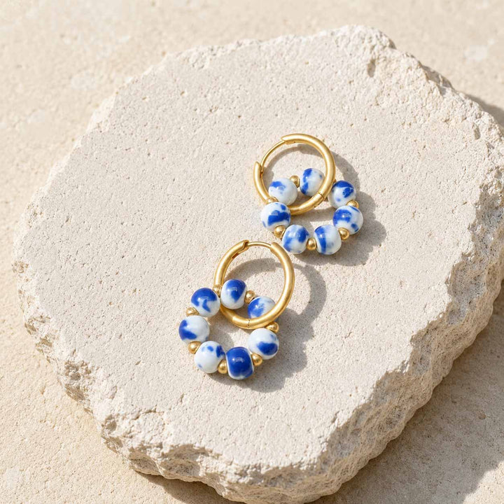 A pair of gold hoop earrings with blue and white porcelain beads, displayed on a textured beige stone against a sandy background.