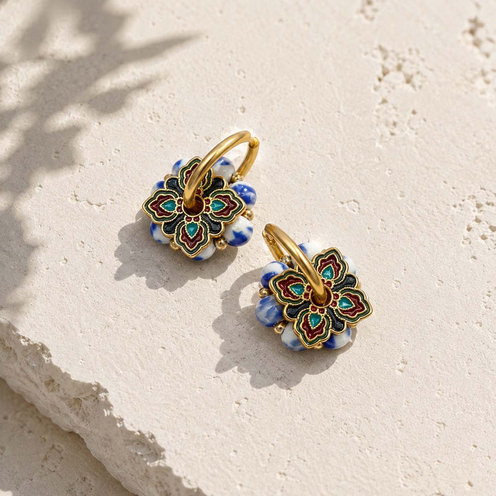 A pair of gold hoop earrings with intricate floral charms featuring blue, red, and green enamel details, displayed on a textured beige stone surface.