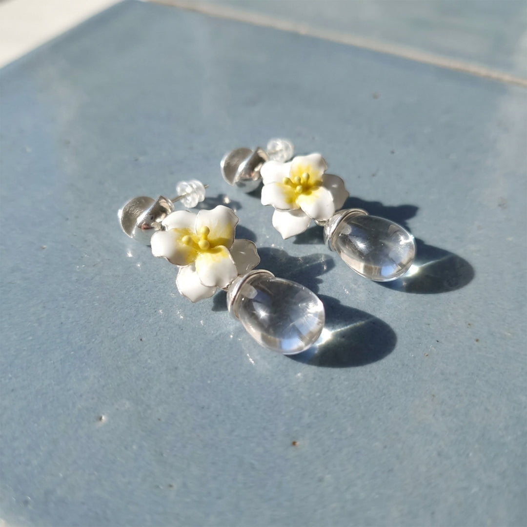 A pair of jasmine clear quartz drop earrings with hand-painted white flowers, resting on a textured blue-gray surface.