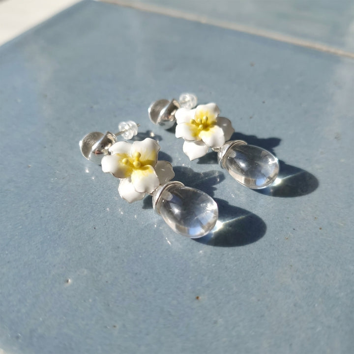 A pair of jasmine clear quartz drop earrings with hand-painted white flowers, resting on a textured blue-gray surface.