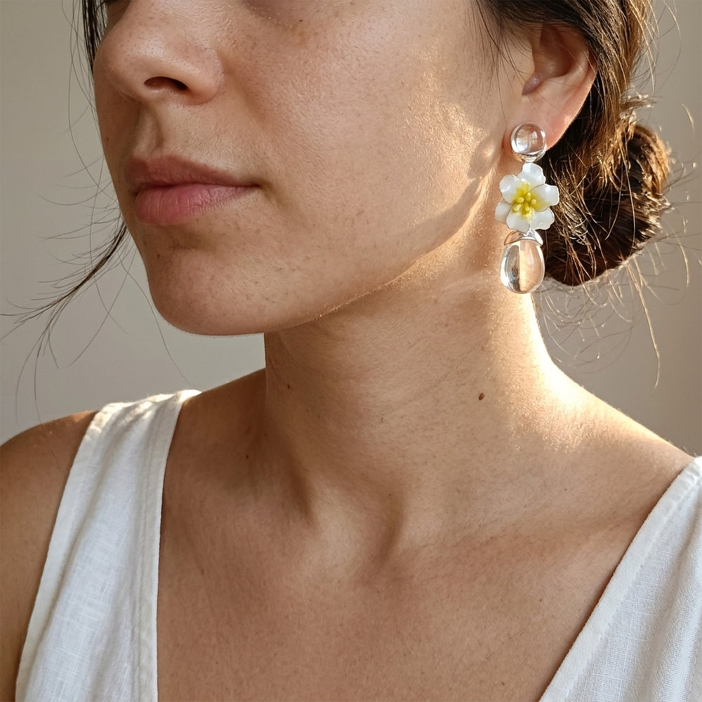 A close-up of a woman wearing a dangling earring with a white flower with a yellow center and clear teardrop charms, paired with a white sleeveless top against a neutral beige background.
