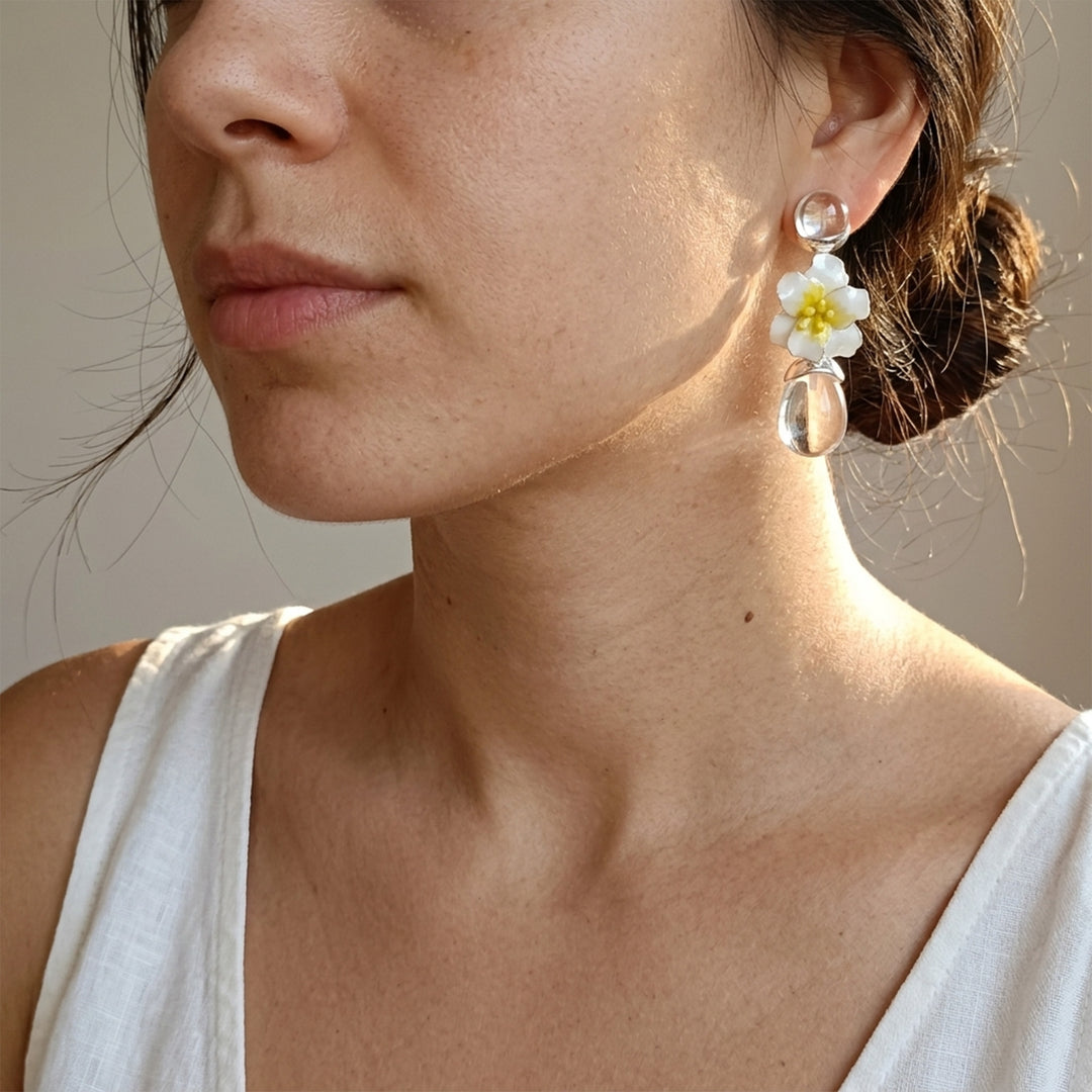 A close-up of a woman wearing a dangling earring with a white flower with a yellow center and clear teardrop charms, paired with a white sleeveless top against a neutral beige background.