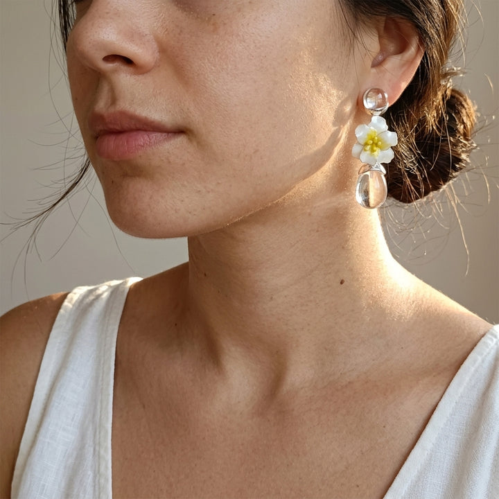 A close-up of a woman wearing a dangling earring with a white flower with a yellow center and clear teardrop charms, paired with a white sleeveless top against a neutral beige background.