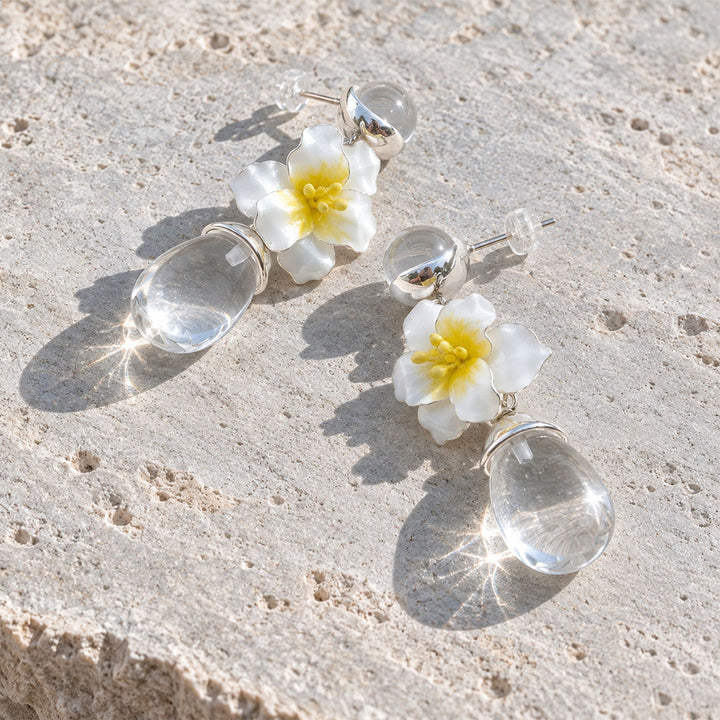 Hand-painted jasmine flower earrings with natural clear quartz drops resting on a textured sandy surface.