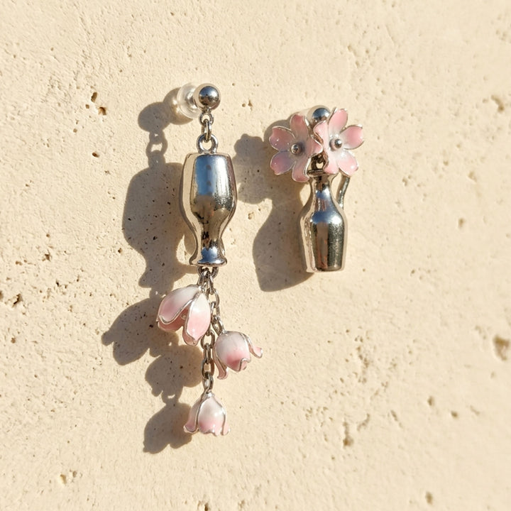 A pair of silver-tone dangle earrings with pink lily of the valley flower details, displayed on a textured beige surface under direct sunlight.