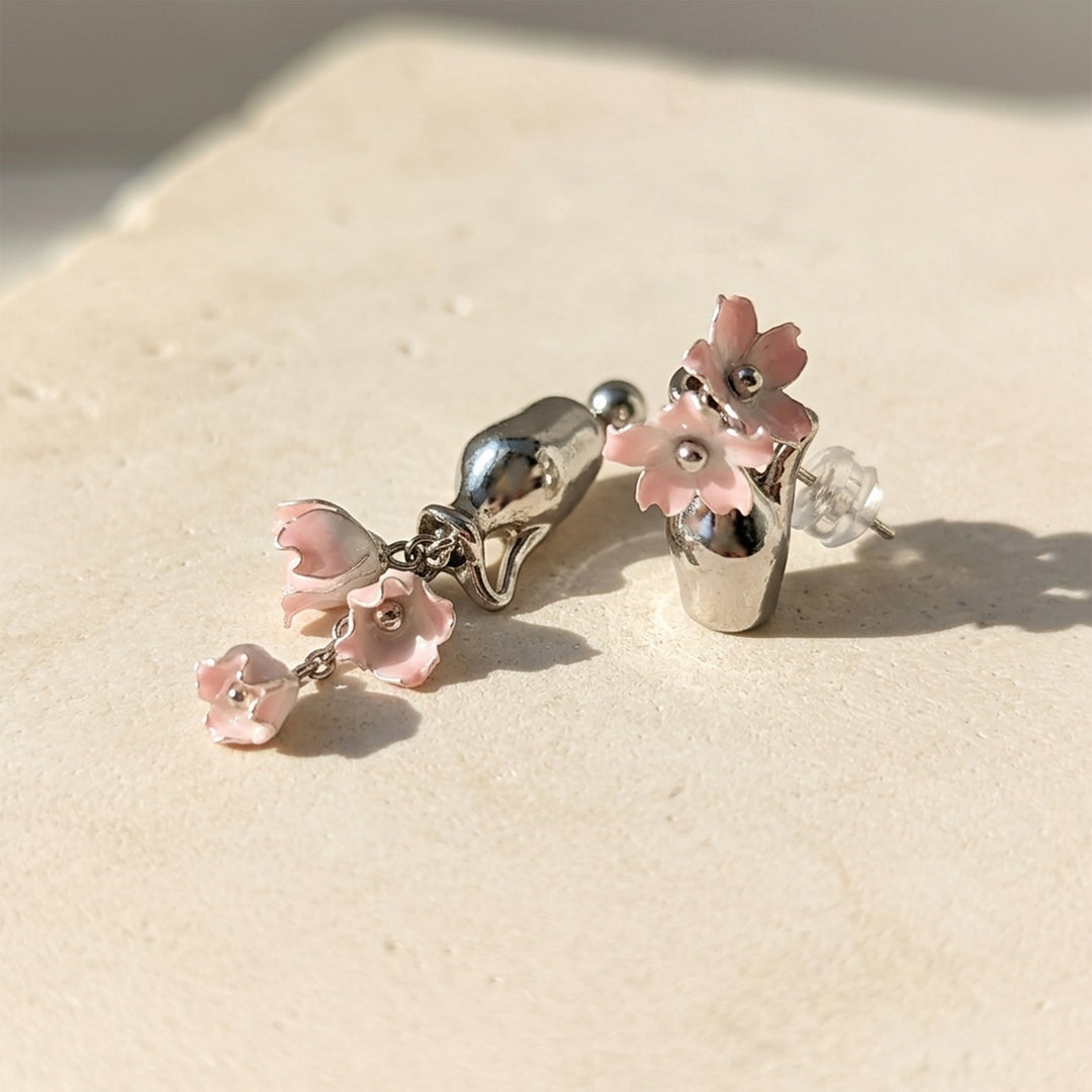 Handcrafted pink Lily of the Valley earrings with silver-plated vase details, resting on a textured beige stone surface under bright natural sunlight.