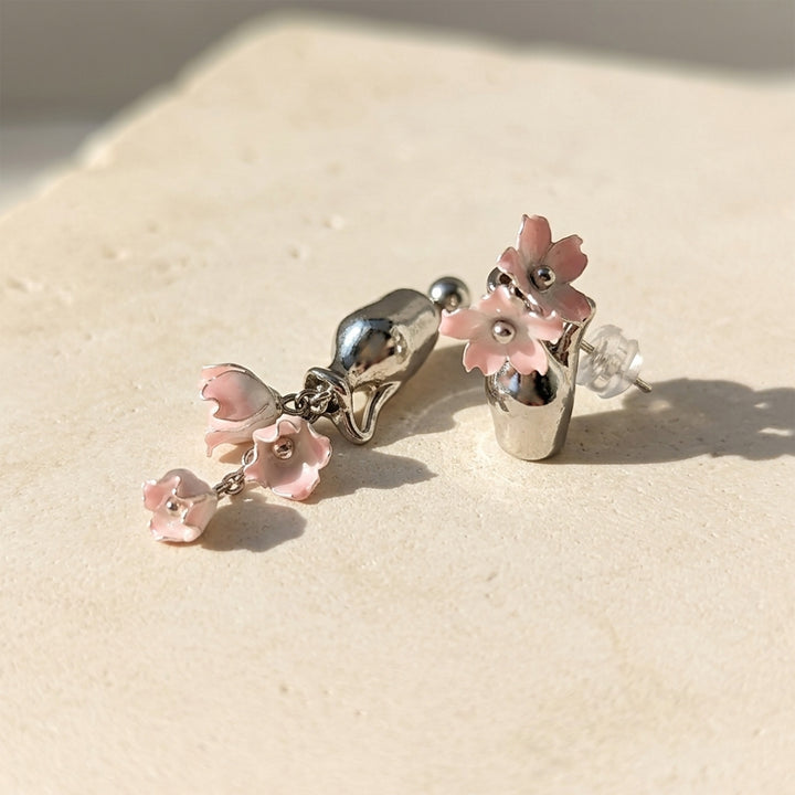 Handcrafted pink Lily of the Valley earrings with silver-plated vase details, resting on a textured beige stone surface under bright natural sunlight.