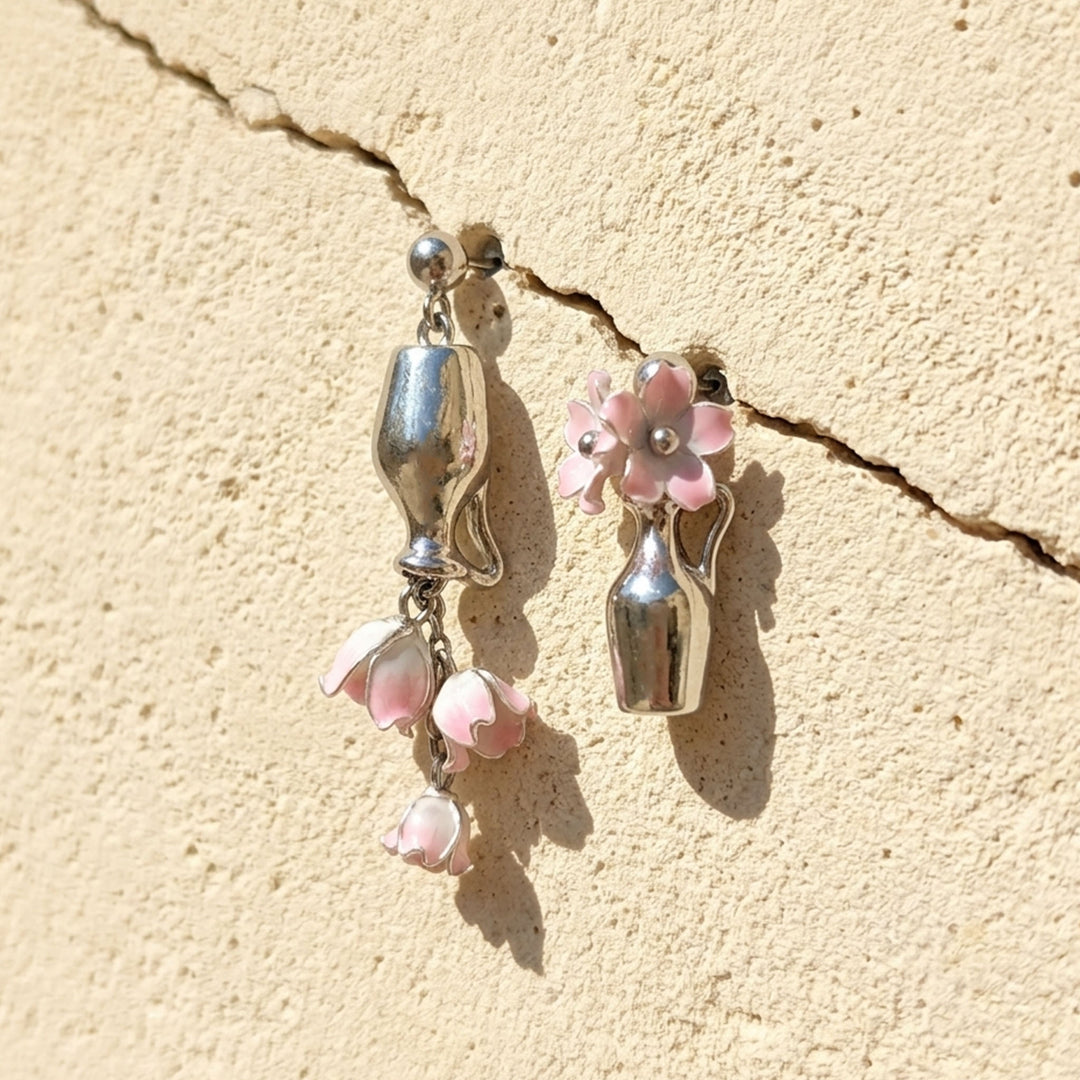 Asymmetric pink Lily of the Valley vase drop earrings with handcrafted flowers and silver-tone details on a sunlit stone wall.