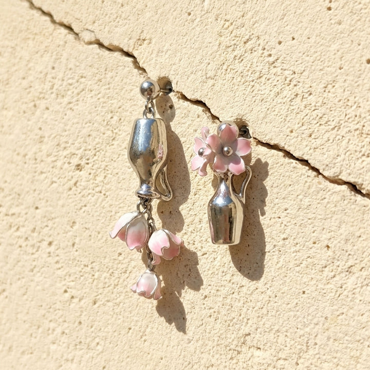 Asymmetric pink Lily of the Valley vase drop earrings with handcrafted flowers and silver-tone details on a sunlit stone wall.