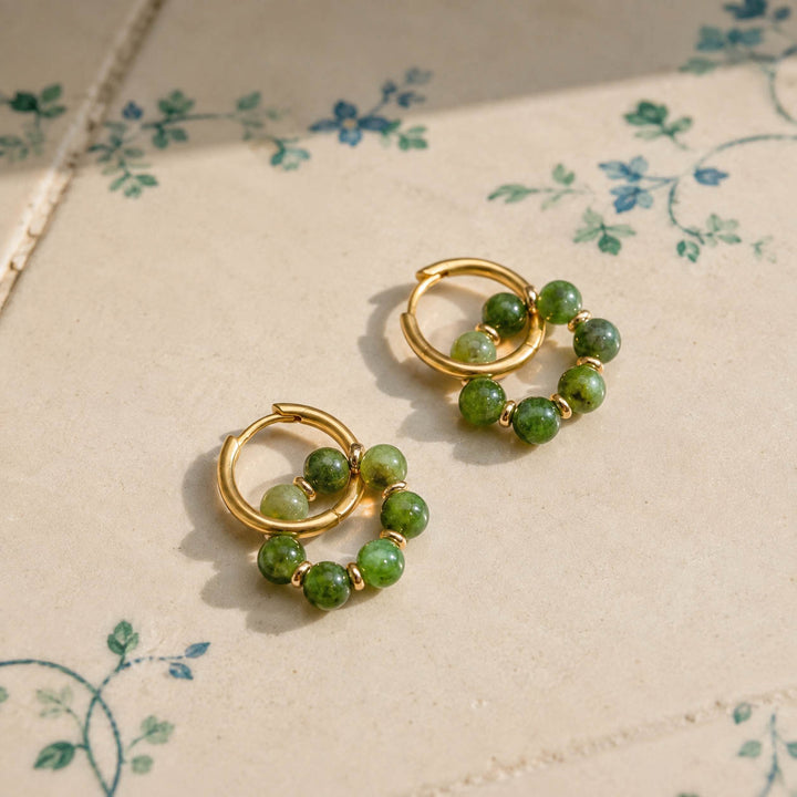 A pair of gold hoop earrings with moss-green Canadian Jade bead clusters, displayed on a beige surface with a delicate blue floral pattern.