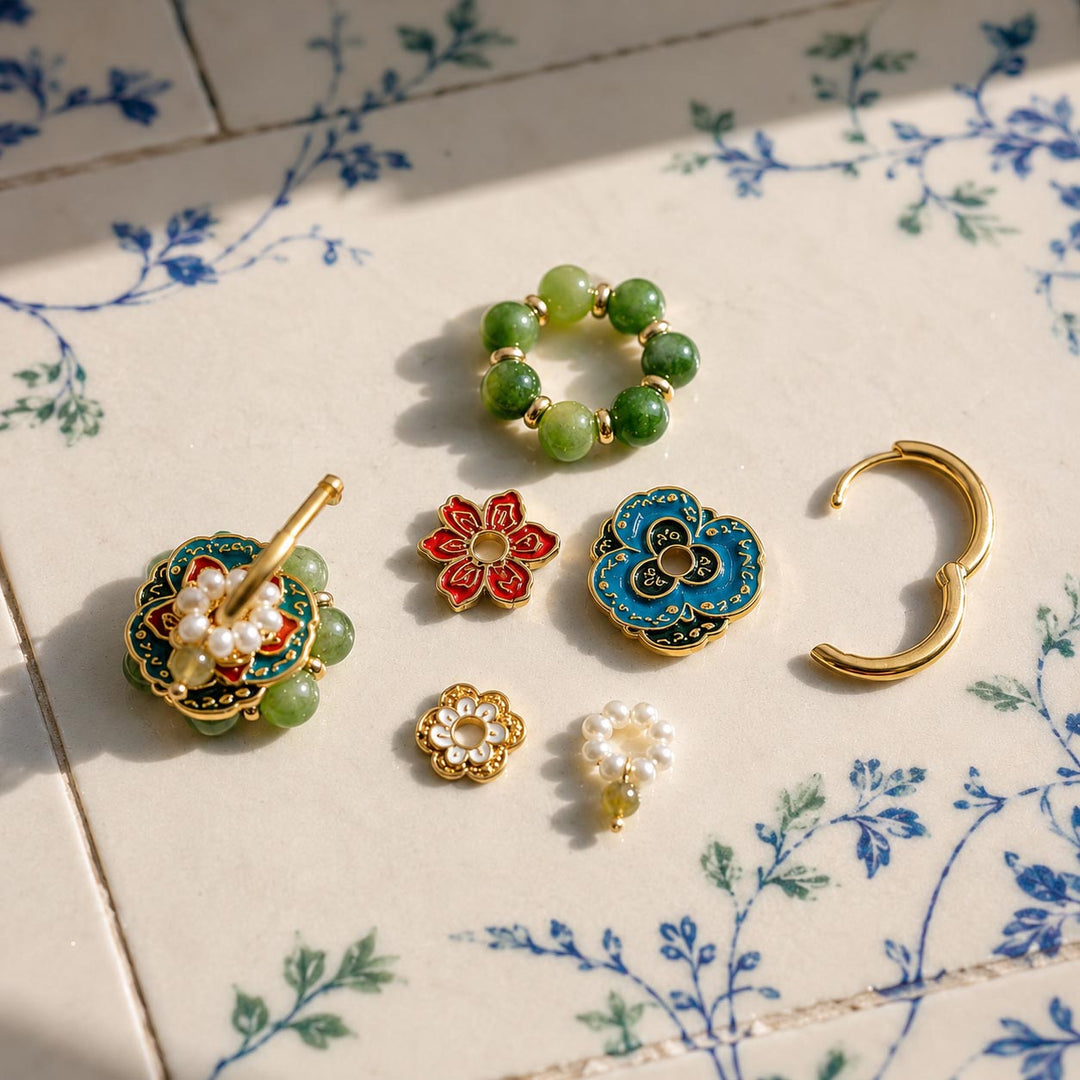 A set of handmade gold-plated jewelry pieces, including enamel flowers, freshwater pearls and green gemstone beads, arranged on a beige ceramic tile with a blue floral pattern.