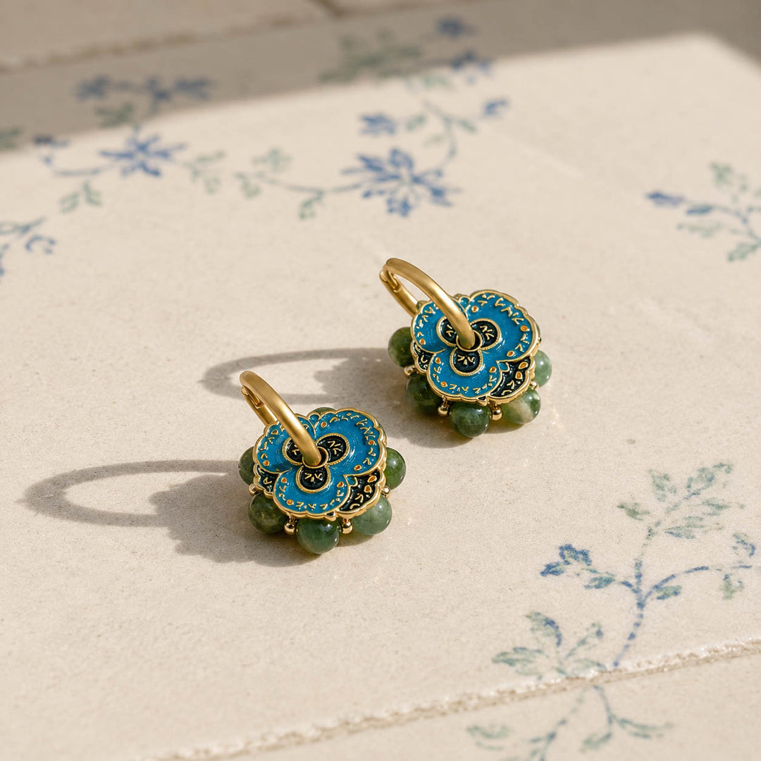 A pair of gold-tone hoop earrings with blue hand-painted enamel flower hoop earrings and moss-green Canadian Jade gemstone beads, resting on a cream-colored surface with a delicate blue floral print.