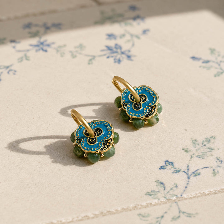A pair of gold-tone hoop earrings with blue hand-painted enamel flower hoop earrings and moss-green Canadian Jade gemstone beads, resting on a cream-colored surface with a delicate blue floral print.